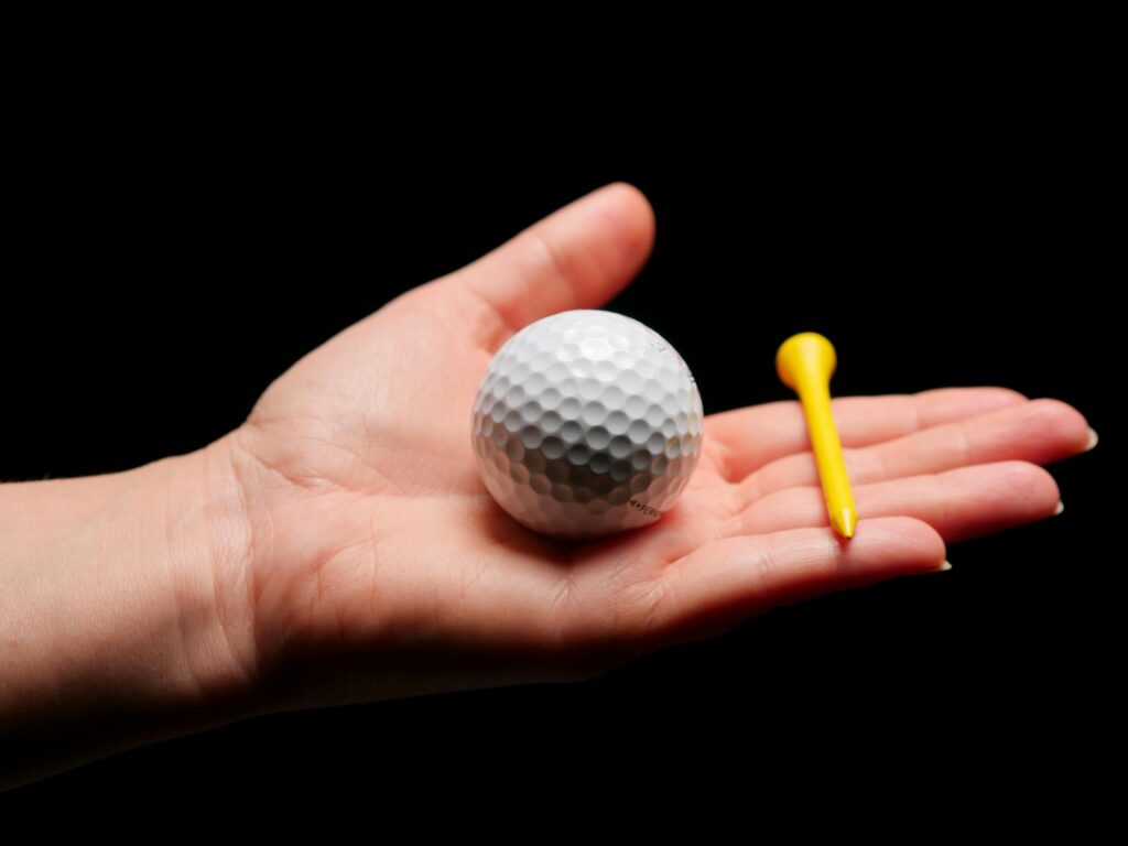 pexels-photo-6573869-6573869 Close-up image of a golf ball and tee in an open palm against a dark background.